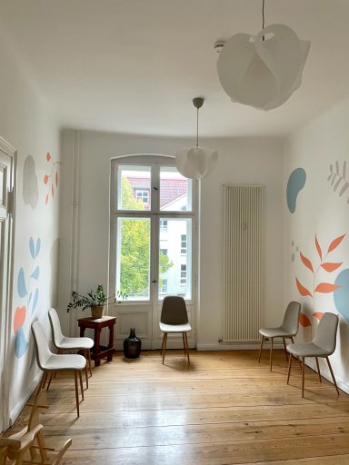 Heller Raum mit Holzboden, weißen Wänden und bunten Wandmalereien, zwei Stühlen und Tisch.